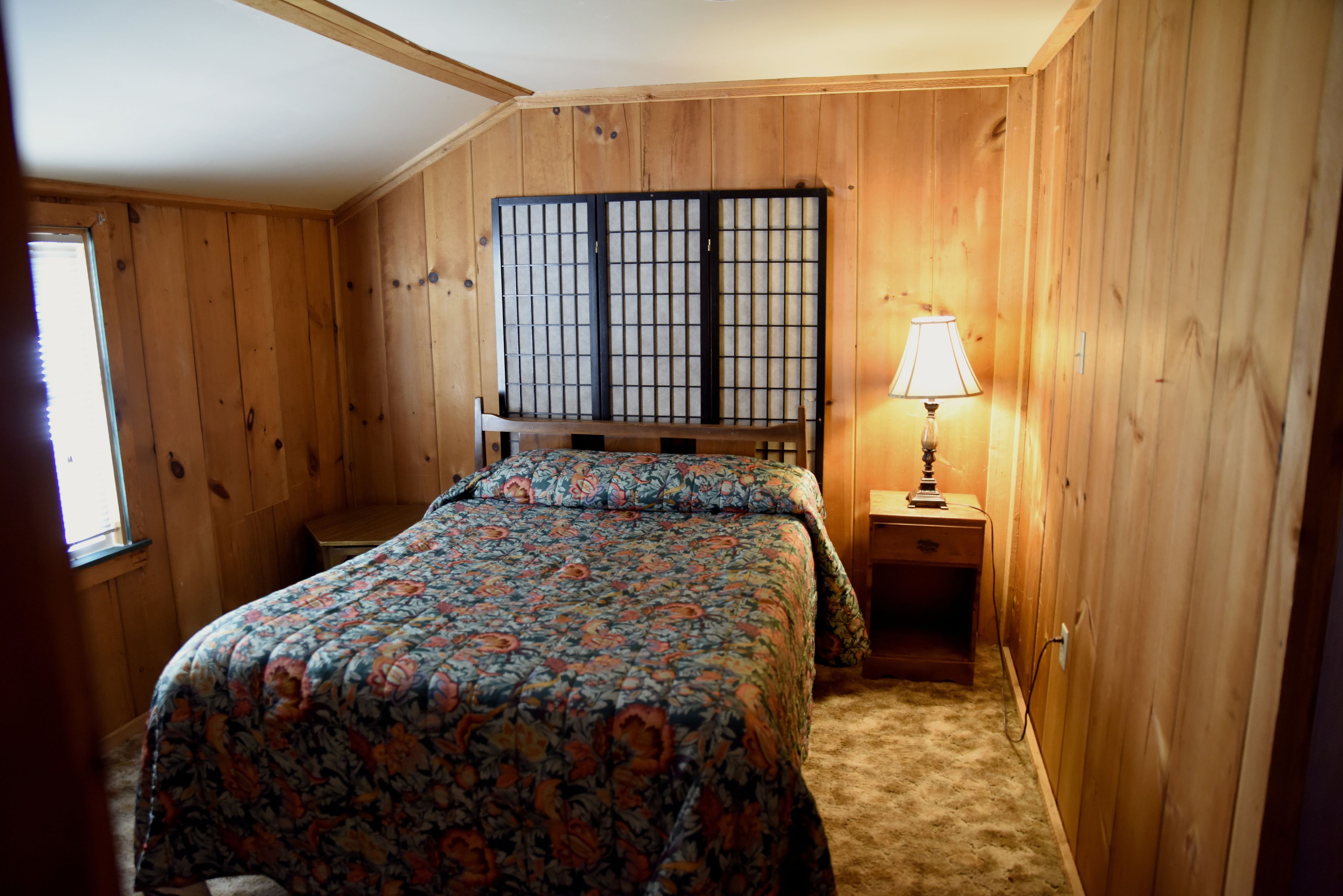 A bedroom with a wooden headboard, a floral bedspread, a bedside lamp, and a window.