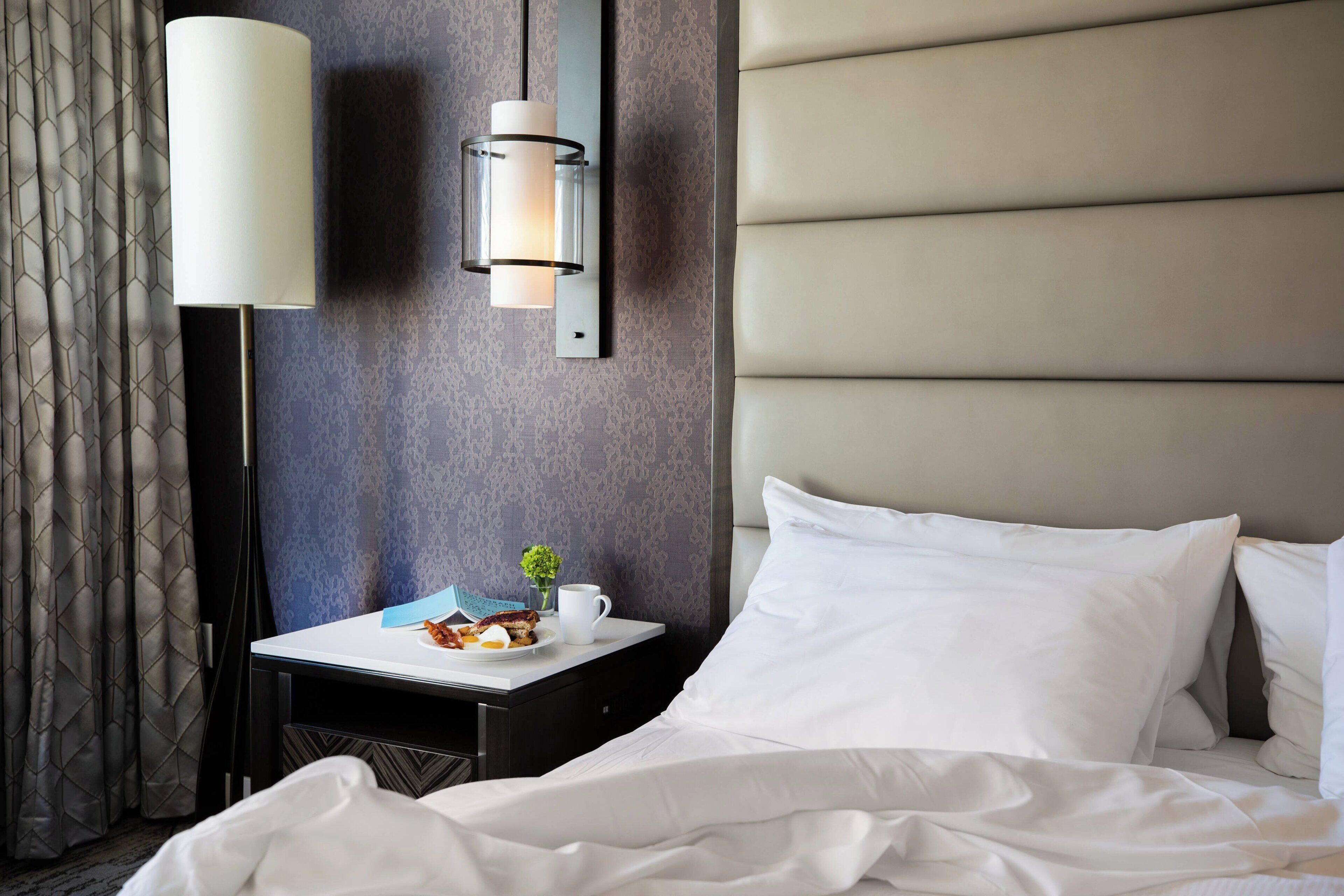 A hotel room with a bed, a bedside table with a lamp, a small table with breakfast items, and a wall-mounted light fixture.