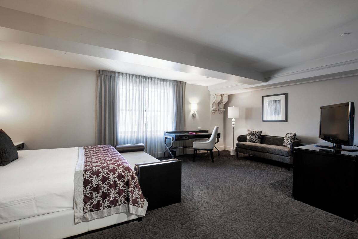 Ein Hotelzimmer mit Bett, Schreibtisch, Sofa, Fernseher und einem großen Fenster mit Vorhängen.
