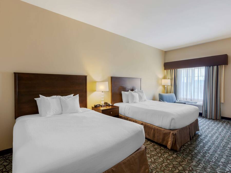 A hotel room with two beds, a nightstand with a lamp, a chair, and a large window with curtains.