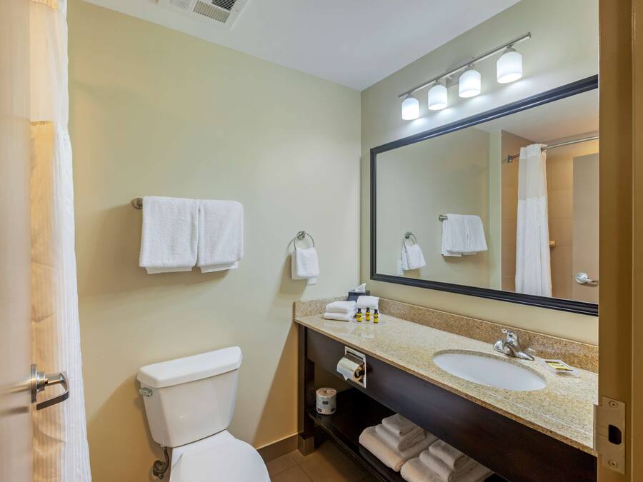 Un baño con inodoro, lavamanos y un espejo grande. Hay toallas colgadas en la pared y un perchero.
