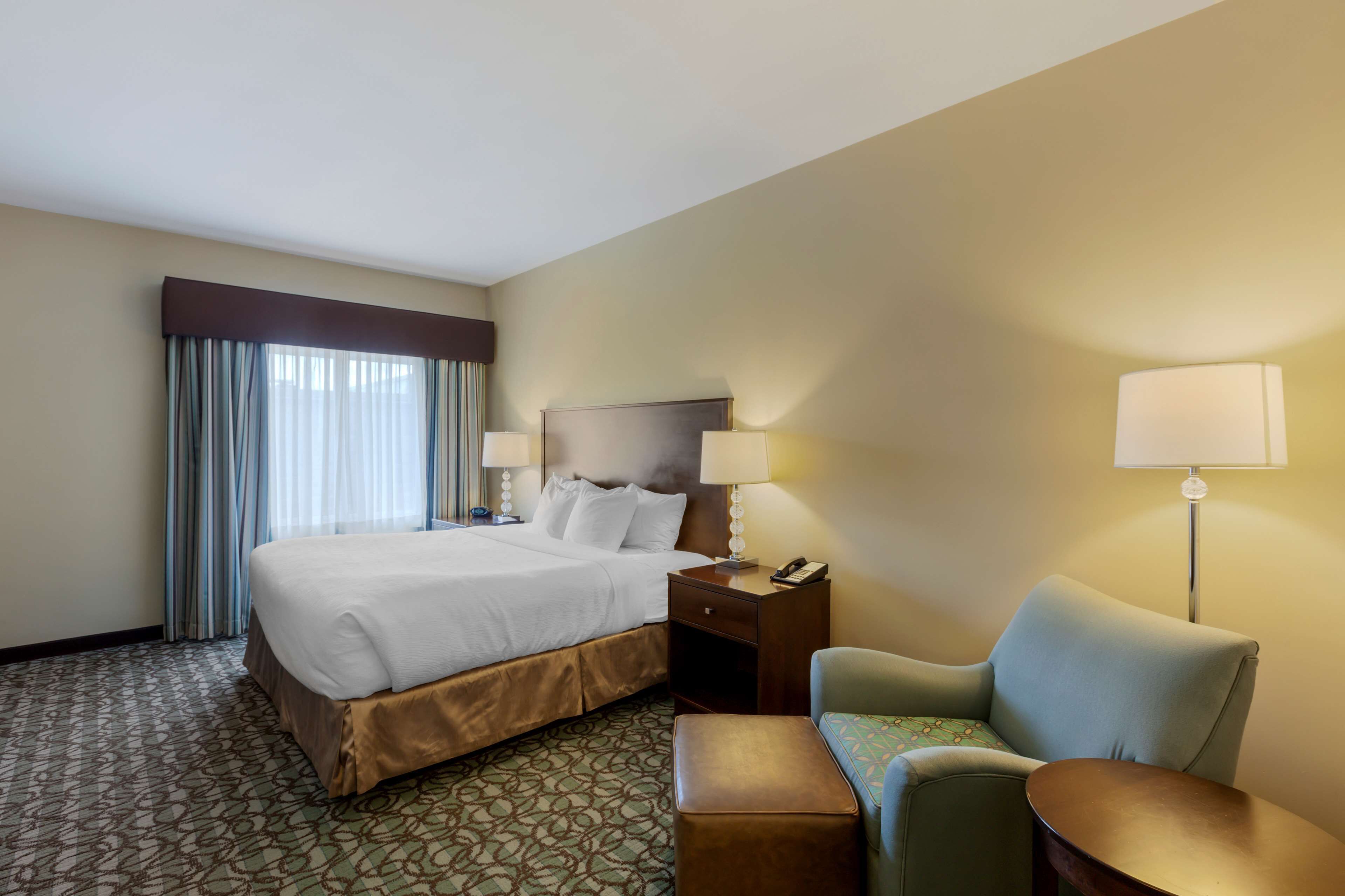 A hotel room with a large bed, a desk with a lamp, a chair, and a patterned carpet.