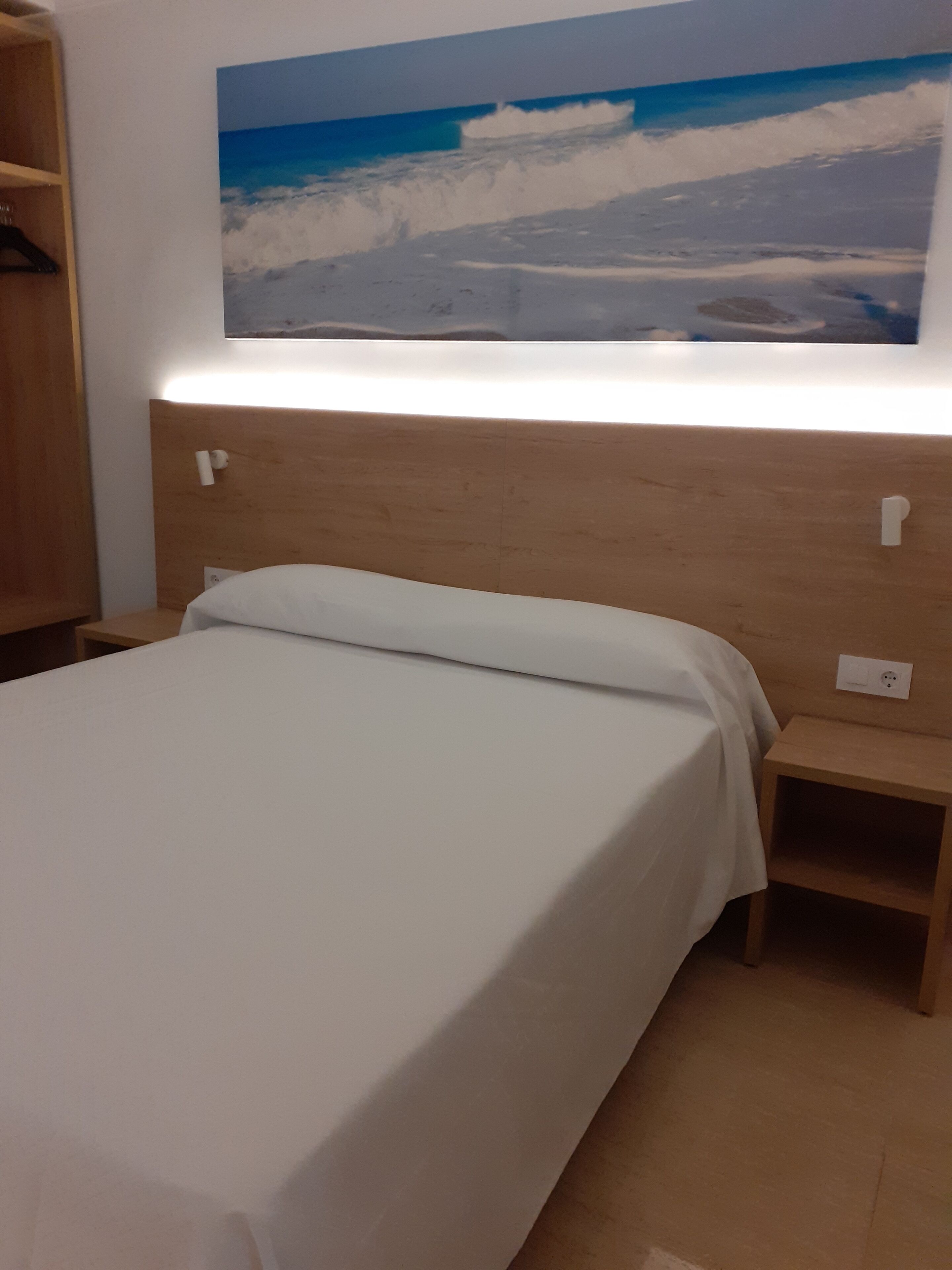 Quarto com cabeceira de madeira, cama com roupa de cama branca, mesa de cabeceira e um quadro pendurado na parede retratando uma cena de praia.