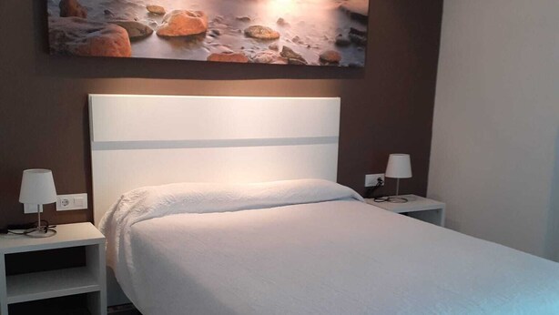 A bedroom with a large bed, two bedside tables with lamps, a nightstand, and a painting of a sunset over rocks on the wall. - Iberflat Hotel Marynton (Benicarlo)