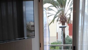 A room with a wooden desk, a television mounted on the wall, and a sliding glass door leading to an outdoor area with a palm tree. - Iberflat Hotel Marynton (Benicarlo)