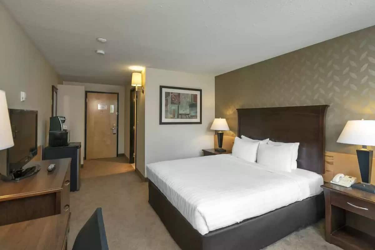 Quarto de hotel com uma cama grande, mesas de cabeceira com luminárias, uma escrivaninha com computador, uma cadeira e uma porta que dá para outro cômodo.