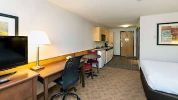 Ett hotellrum med en säng, ett skrivbord med en stol, en tv, en lampa, ett litet pentry och en dörr som leder till ett annat rum.