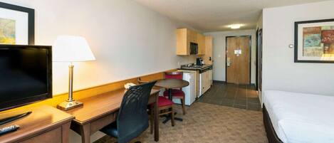 A hotel room with a bed, a desk with a chair, a television, a lamp, a small kitchenette, and a door leading to another room.