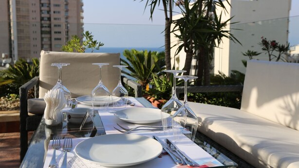Tavola imbandita per la cena con bicchieri trasparenti, piatti e posate, affacciata su una vista costiera con edifici e palme.