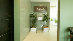 A reception area with a sign reading "Pacifica Cafe," two potted plants, a reception desk, and a glass door leading to a seating area. - Grand Pacific Hotel PWTC (Kuala Lumpur)