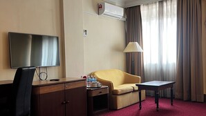 A hotel room with a red carpet, a sofa, a desk with a TV, a lamp, and an air conditioning unit. - Grand Pacific Hotel PWTC (Kuala Lumpur)