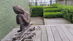 A Buddha statue on a wooden platform with a hedge and a concrete path in the background. - Designer Cottage B&B (Christchurch)