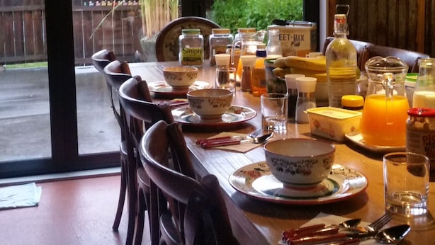 A dining table set with various dishes, glasses, and condiments, with a view of a garden and a wooden fence outside. - Designer Cottage B&B (Christchurch)