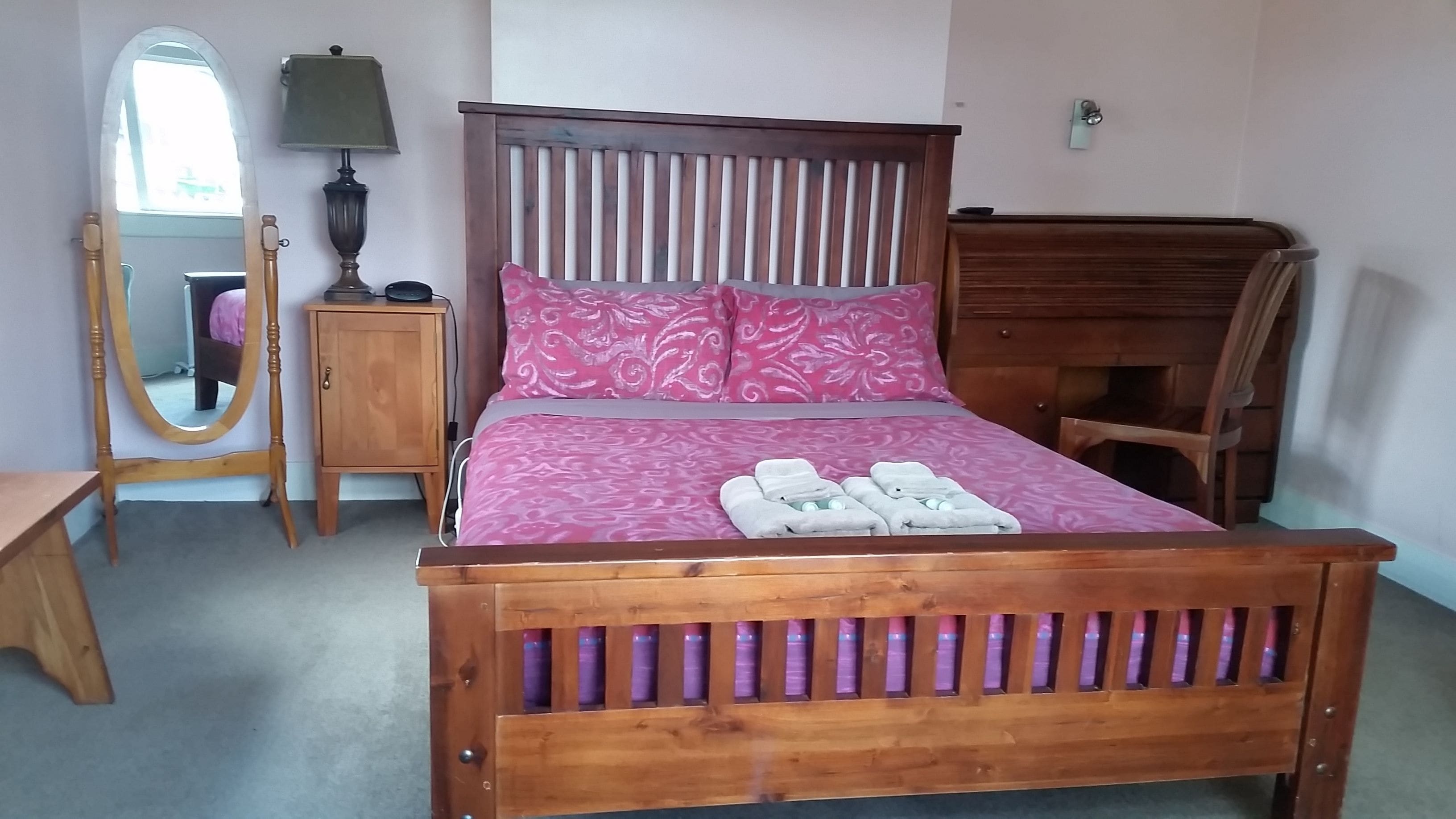 A wooden bed with a pink patterned bedspread and pillows, a wooden nightstand with a lamp, a wooden chair, and a wooden desk.