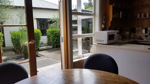 A kitchen with a white microwave on the counter, a wooden table, and a view of a house and garden through the window. - Designer Cottage B&B (Christchurch)