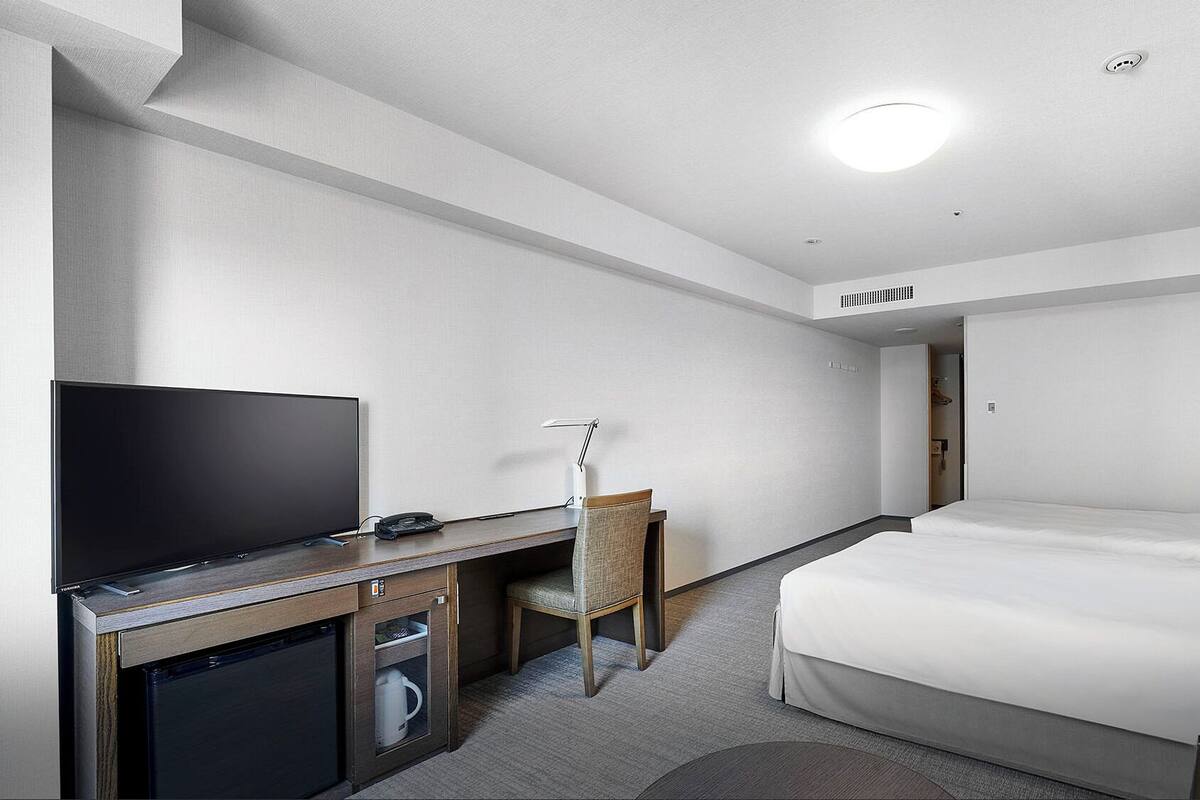 A hotel room with a bed, a desk with a TV, a chair, and a small kitchenette.