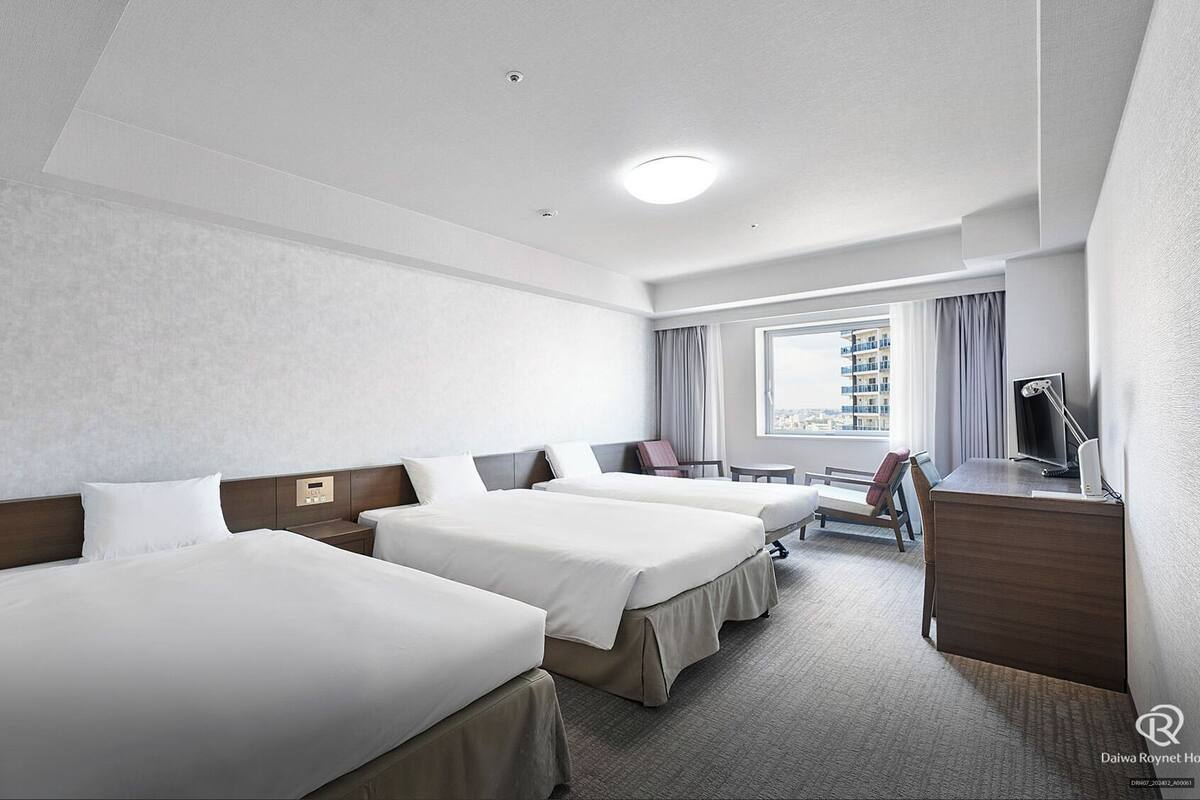 A hotel room with three single beds, a desk with a computer, a chair, and a window with a view of buildings.