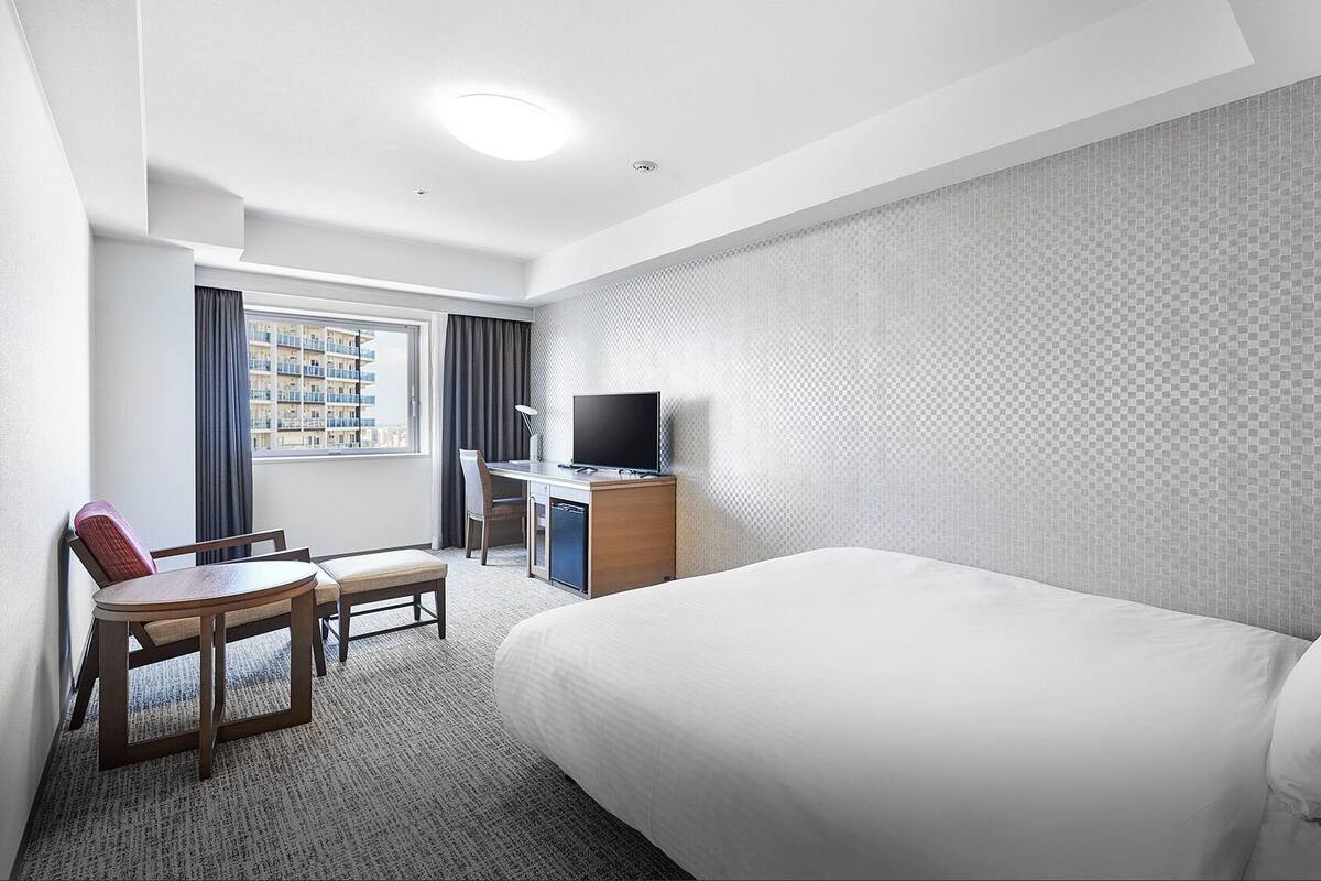 A hotel room with a bed, a desk with a TV, a chair, a small table, and a window with curtains.