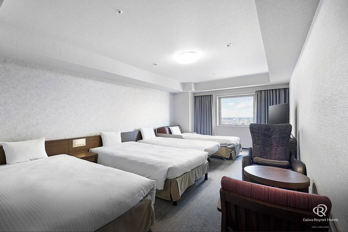 A hotel room with three beds, a desk, a chair, and a view of the city.