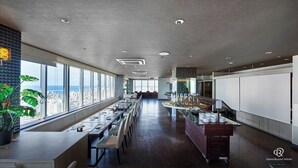 A modern hotel dining area with long tables, a buffet station, and large windows offering a city view.