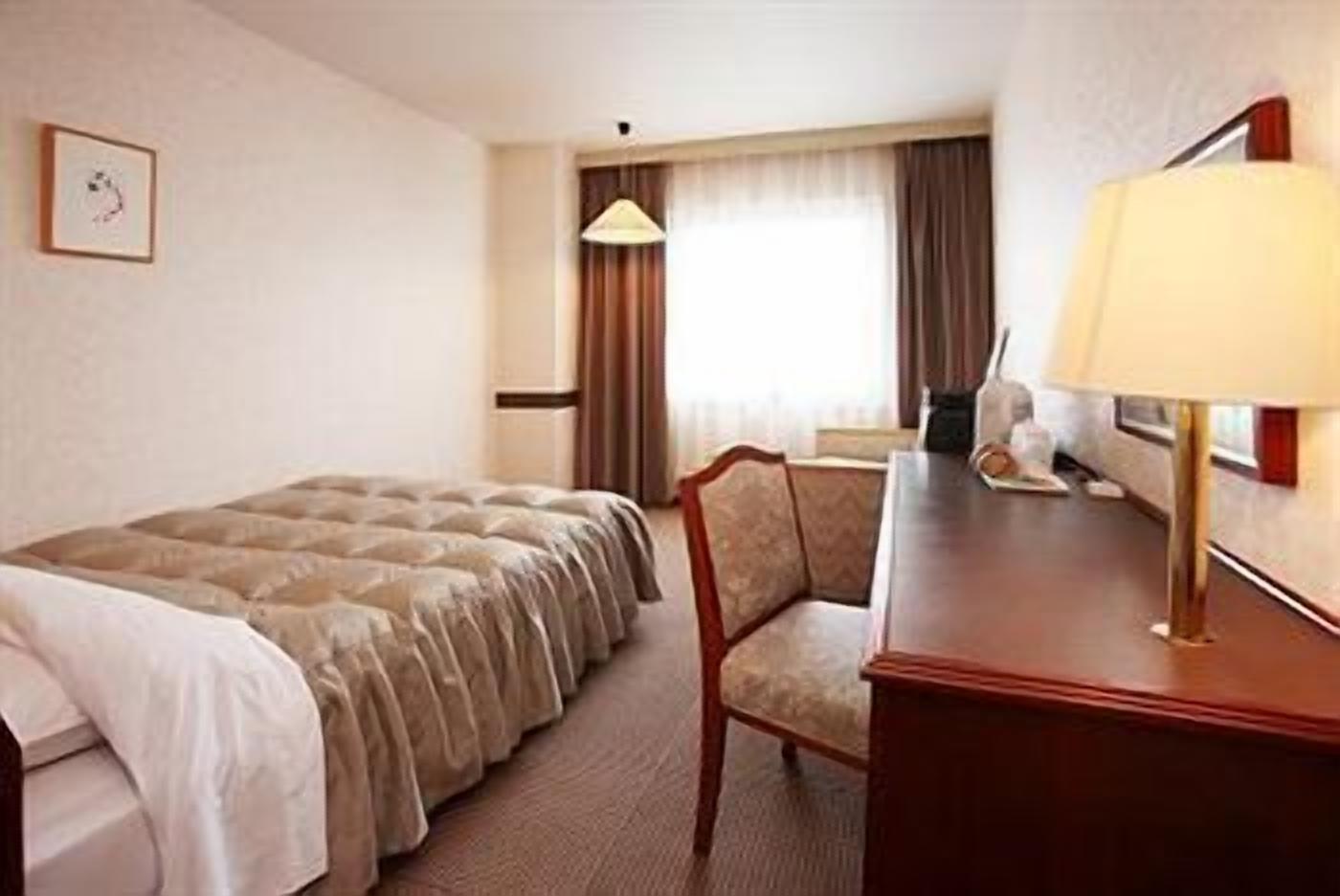 Een hotelkamer met een bed, een bureau, een stoel, een lamp en een raam met gordijnen.