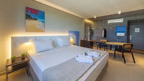 Ein modernes Hotelzimmer mit einem großen Bett, einem Schreibtisch und einem Sessel. An der Wand hängt ein Plakat und auf dem Nachttisch steht eine kleine Pflanze.