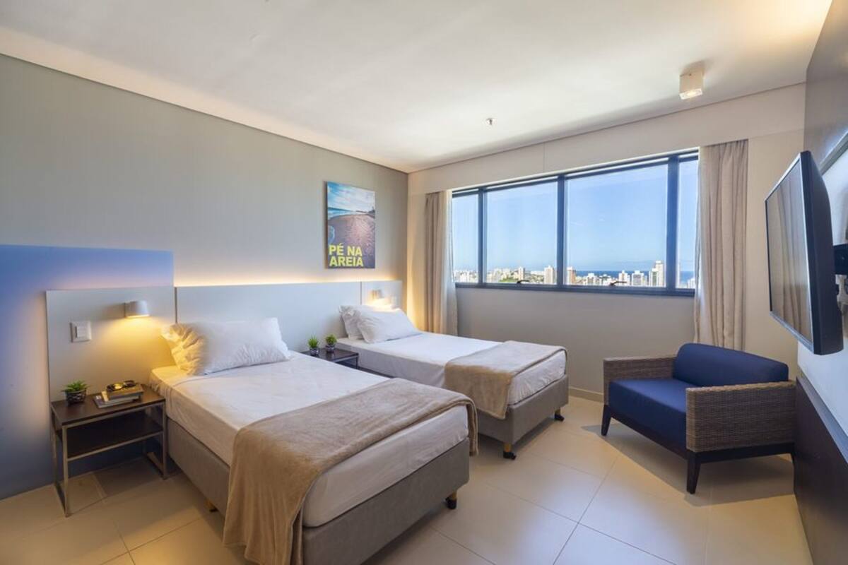 A hotel room with two beds, a TV, a window with a city view, and a chair.