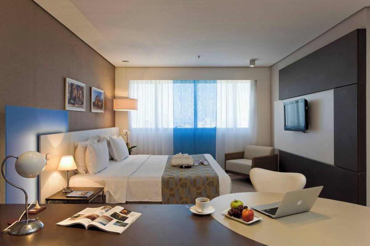 A hotel room with a bed, bedside table with a lamp, a desk with a laptop, a chair, a TV mounted on the wall, and a window with curtains.