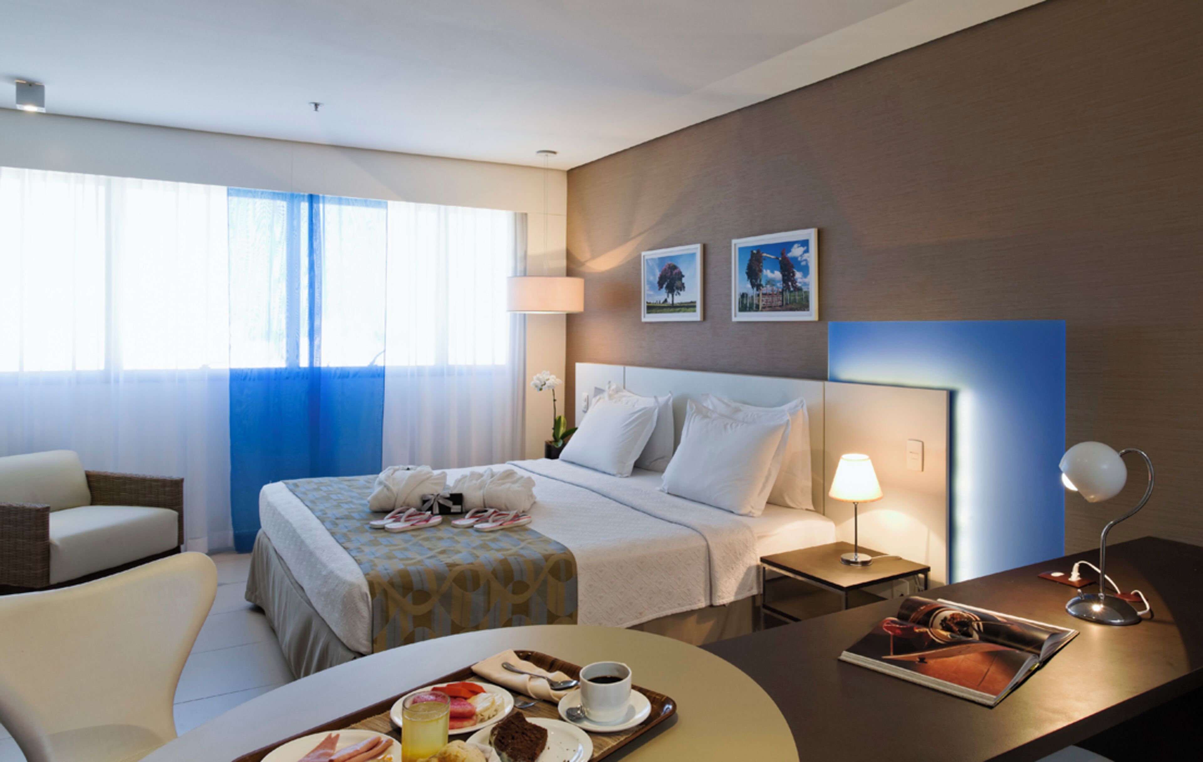 A hotel room with a bed, a desk with a lamp, a chair, a dining table with food, and a window with curtains.