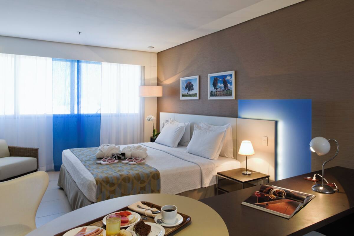 A hotel room with a bed, a desk with a lamp, a chair, a dining table with food, and a window with curtains.