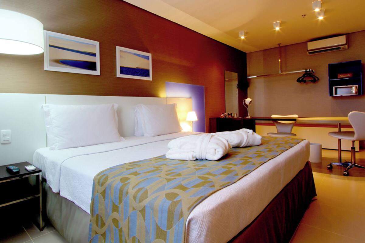 A hotel room with a bed, bedside table, lamp, and a desk with a chair.