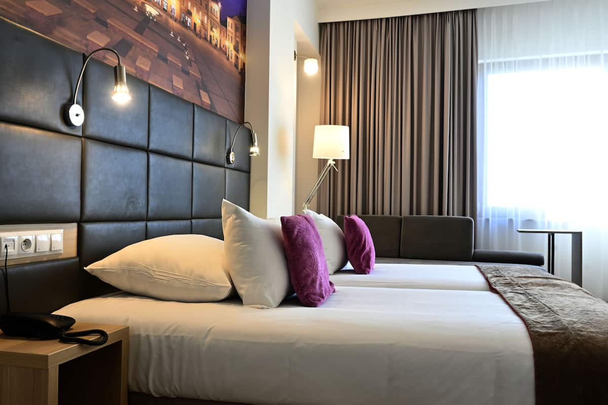 Une chambre d’hôtel avec un grand lit, une tête de lit dotée d’un éclairage intégré, une table de chevet avec un téléphone et une fenêtre avec des rideaux.