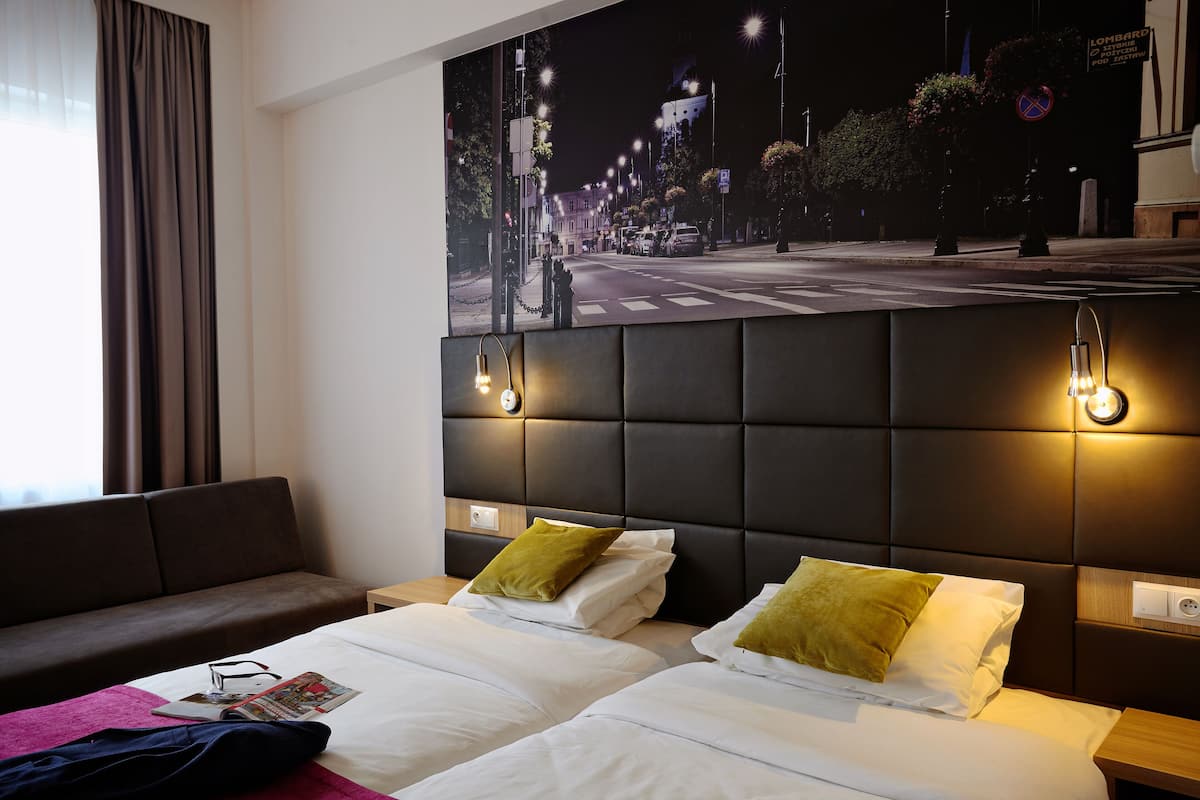 Une chambre d’hôtel avec deux lits, un canapé, une grande affiche représentant un paysage urbain et une tête de lit moderne dotée d’un éclairage intégré.