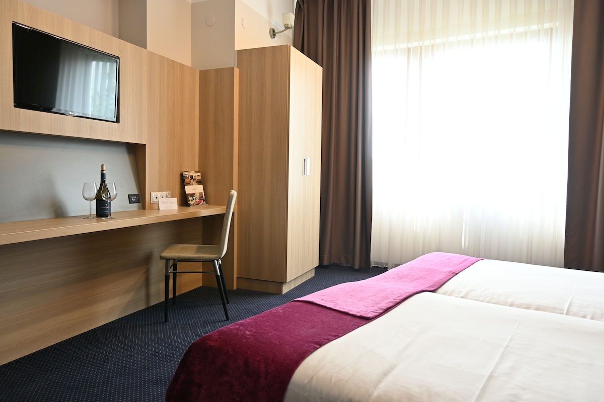 A hotel room with a bed, a desk with a chair, a TV mounted on the wall, and a window with curtains.