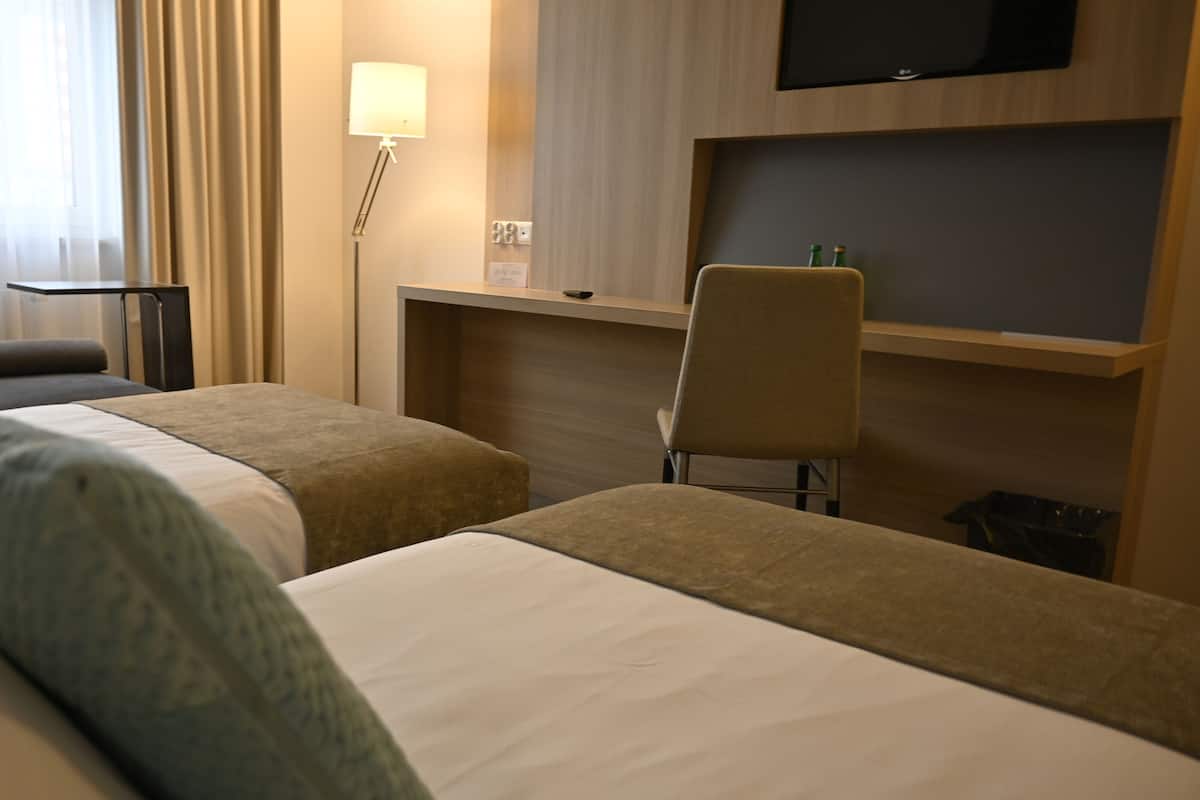 Une chambre d’hôtel avec deux lits simples, un bureau, une chaise, une lampe et un téléviseur à écran plat fixé au mur.