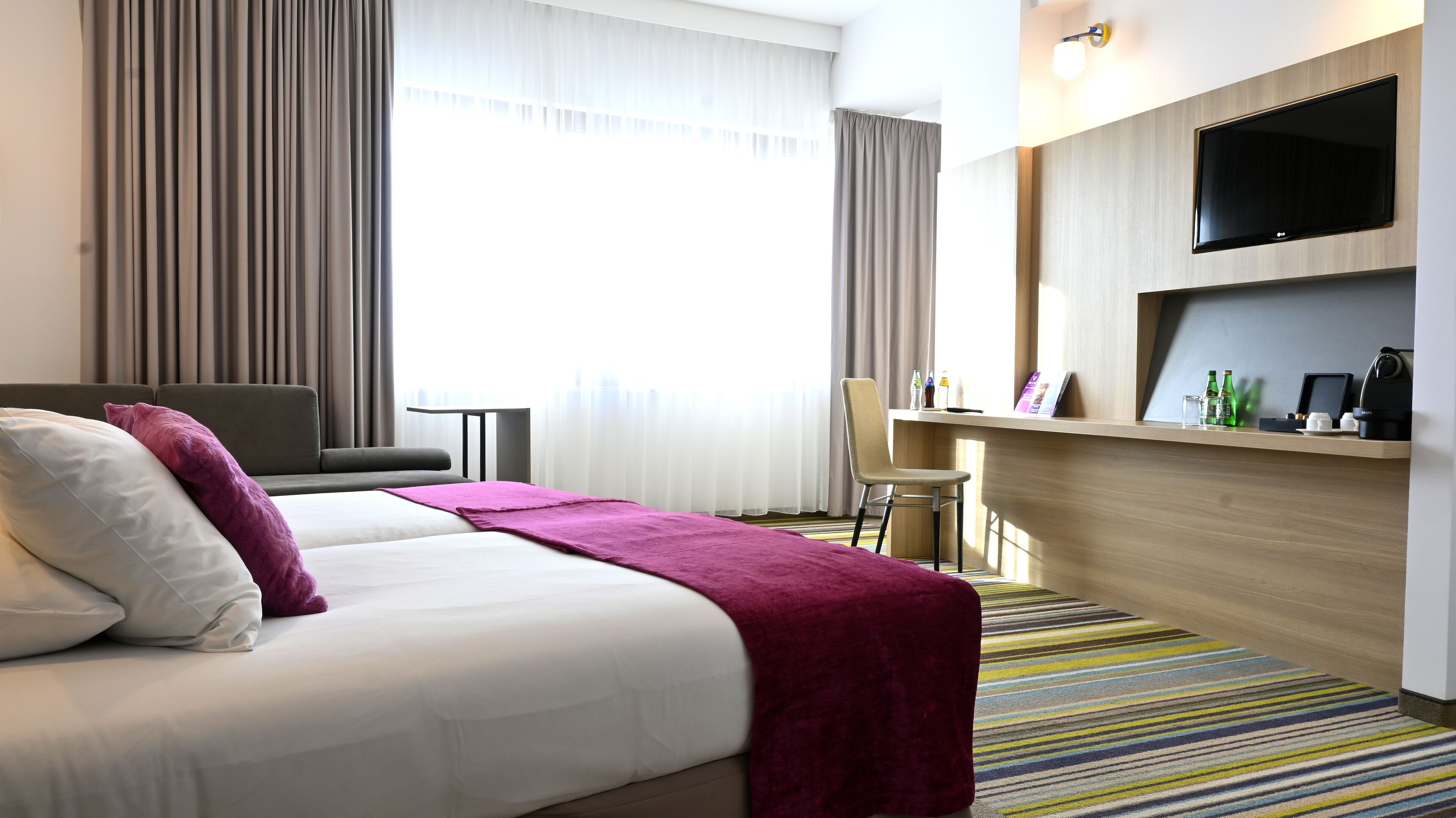 A hotel room with a bed, a desk, a chair, a TV, and a window with curtains.