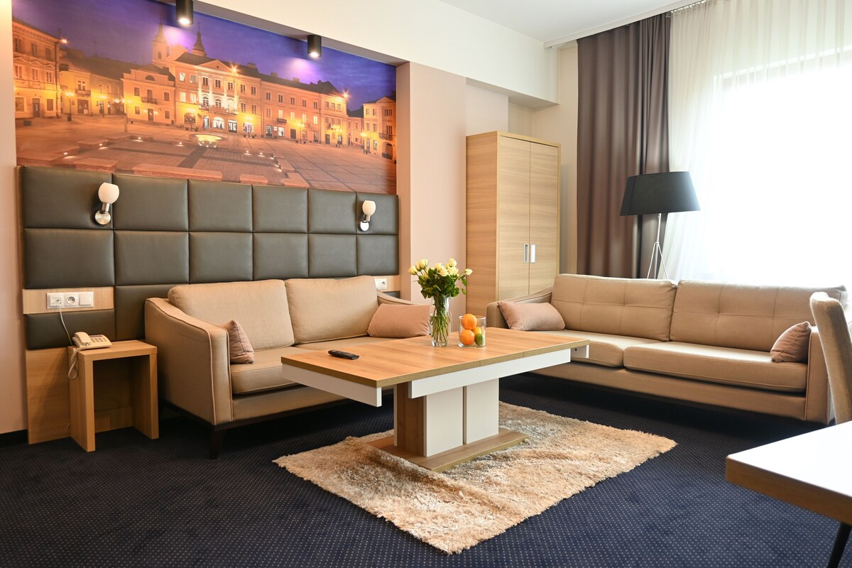 Une chambre d’hôtel moderne avec un grand lit, un canapé, une table basse et un tableau représentant un paysage urbain.