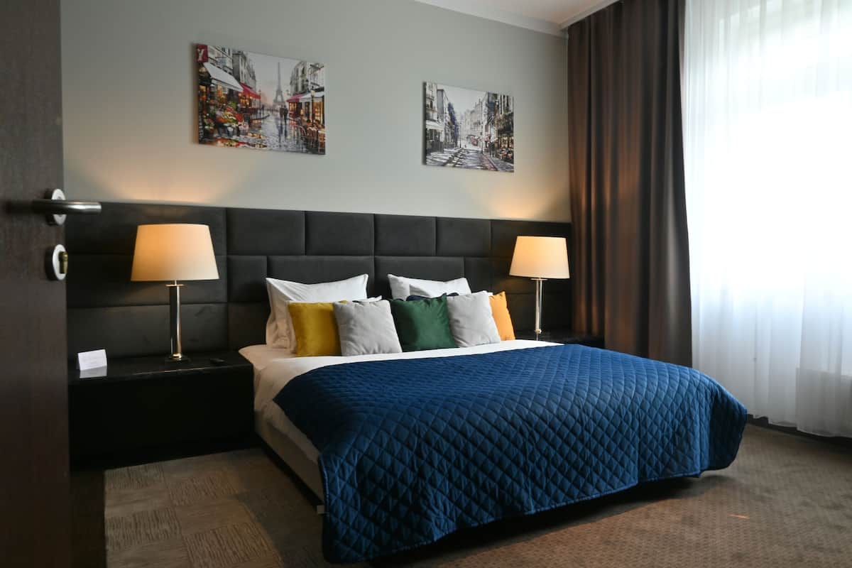 Une chambre d’hôtel avec un grand lit, deux tables de chevet avec des lampes, une tête de lit sombre et deux œuvres d’art mural représentant des paysages urbains.