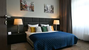 Hotellihuone, jossa on suuri sänky, kaksi yöpöytää valaisimineen, tumma pääty sekä kaksi seinätaideteosta, joissa on kuvattu kaupunkimaisemia.