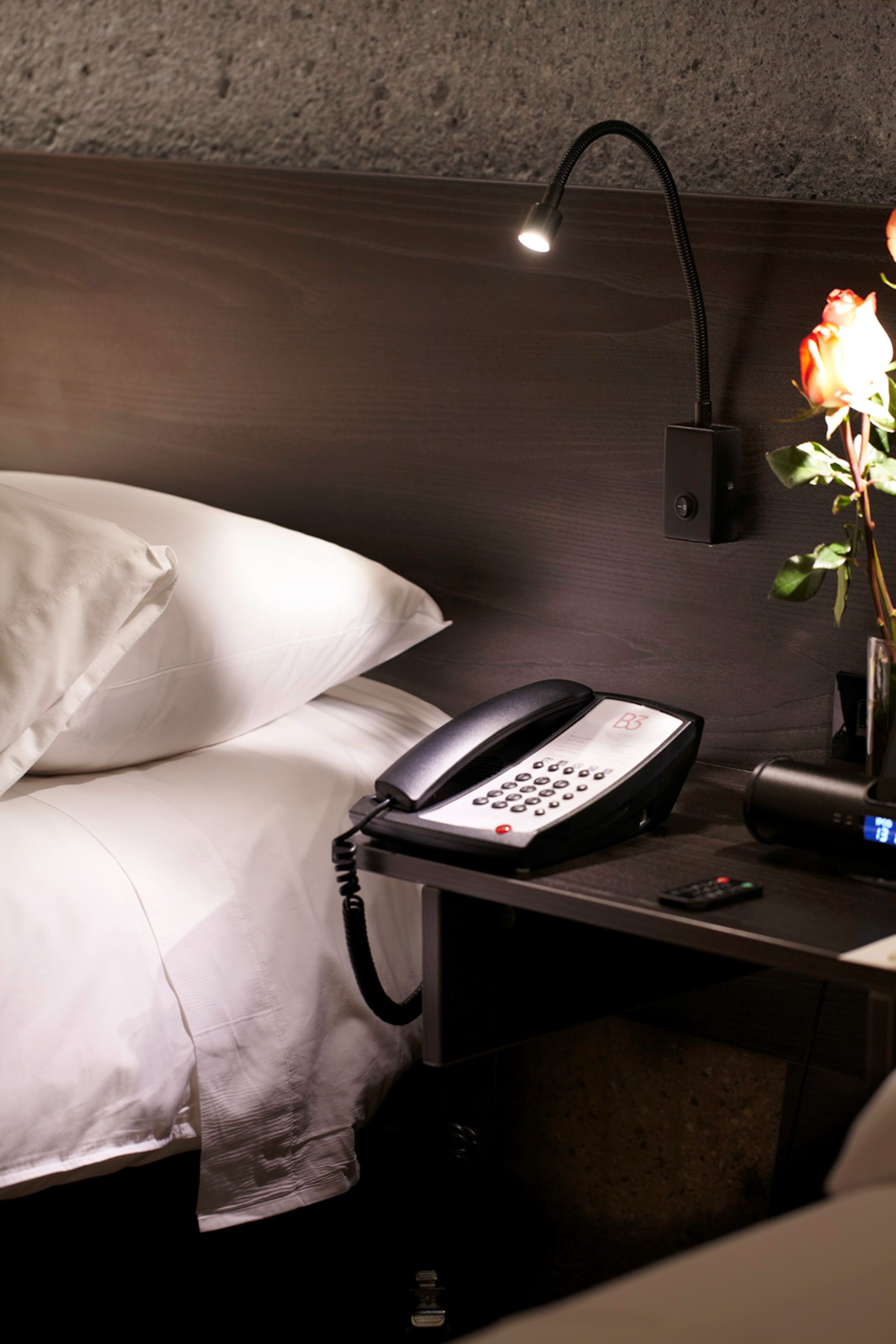 A hotel room with a bed, a bedside telephone, a lamp, and a vase with a flower.