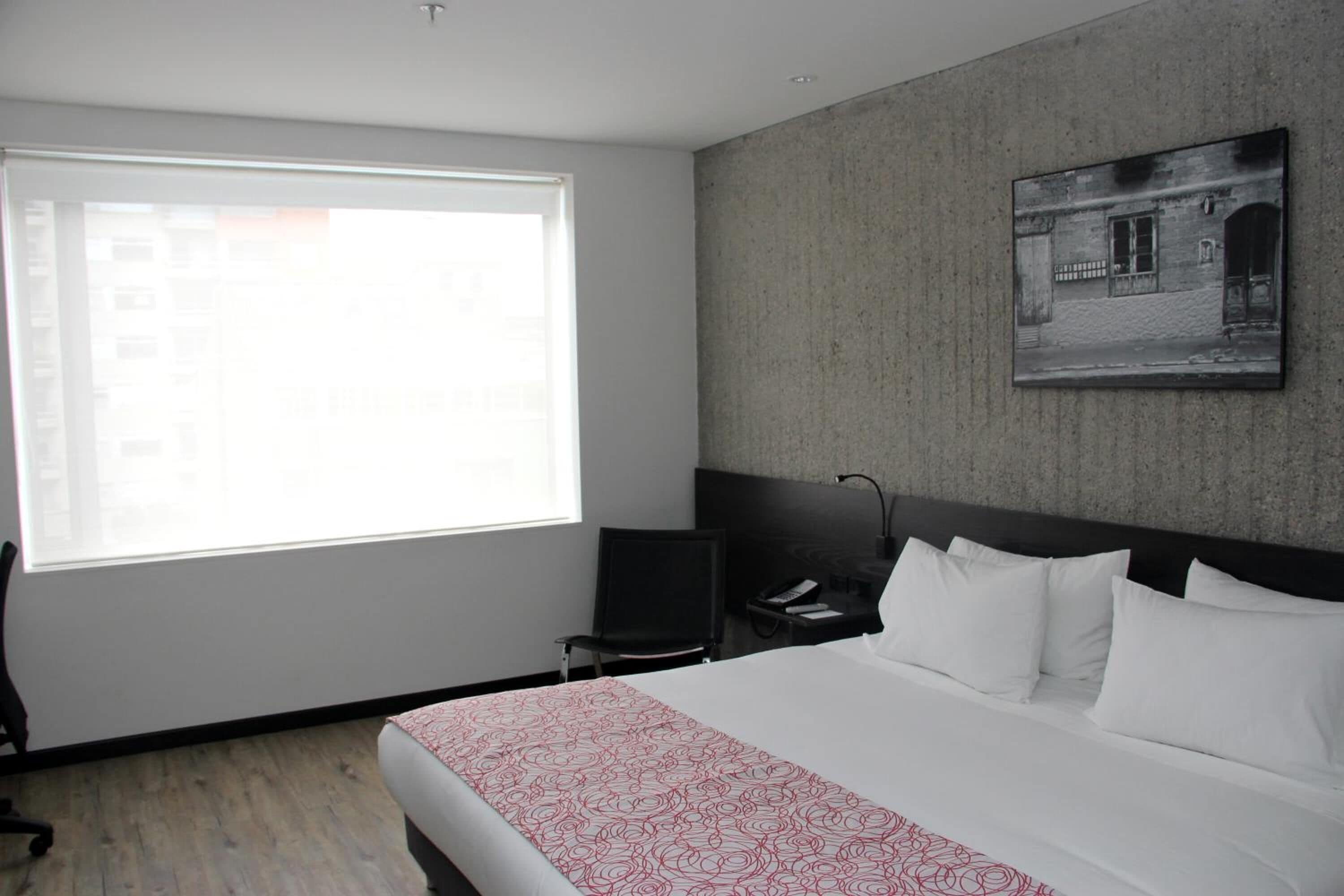 Een hotelkamer met een groot raam, een bed met wit en rode beddengoed, een bureau met een stoel en een ingelijste foto aan de muur.
