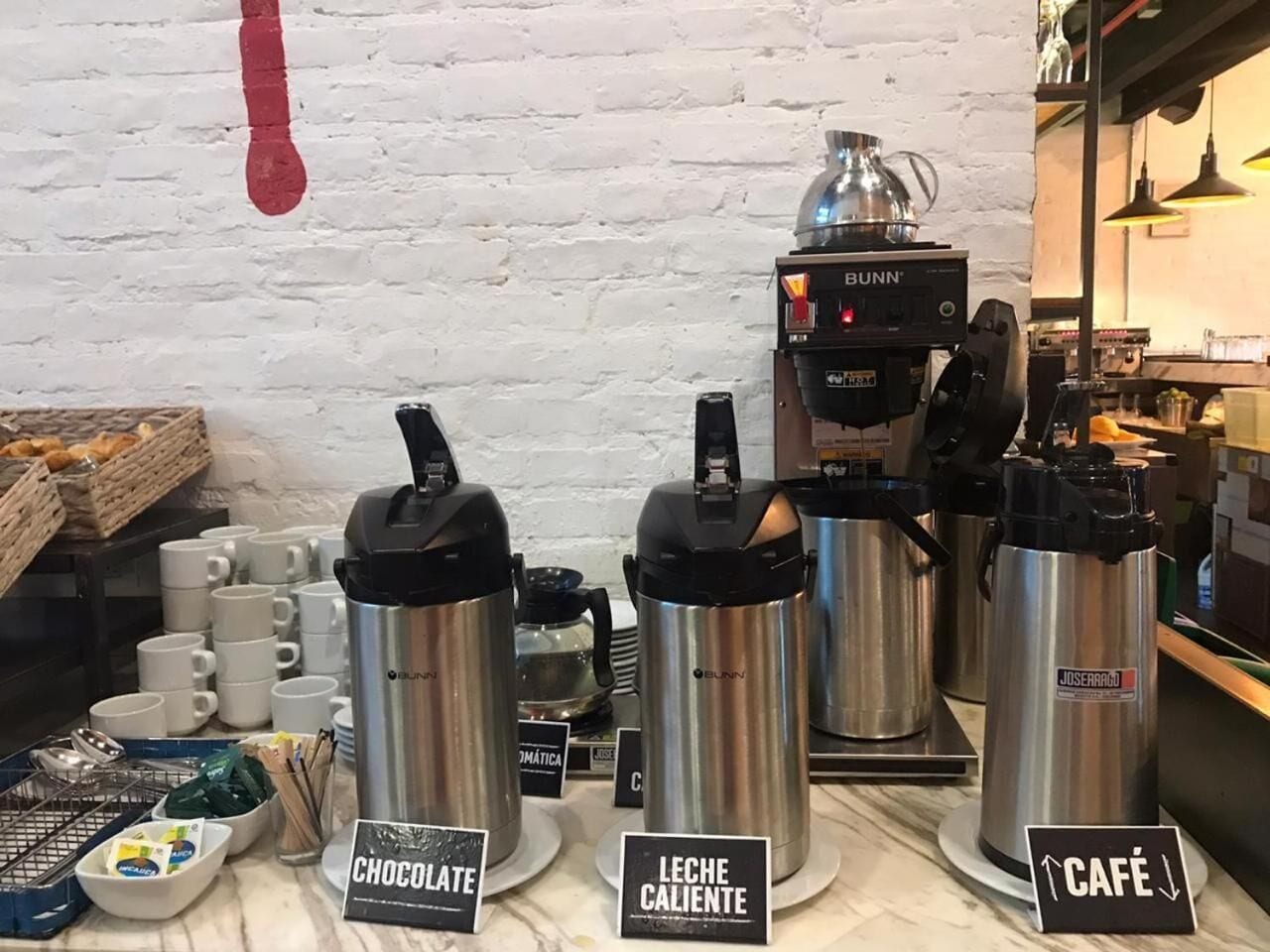 A coffee shop with a Bunn espresso machine, thermos cups labeled 'Chocolate' and 'Café', and a 'Leche Caliente' sign.