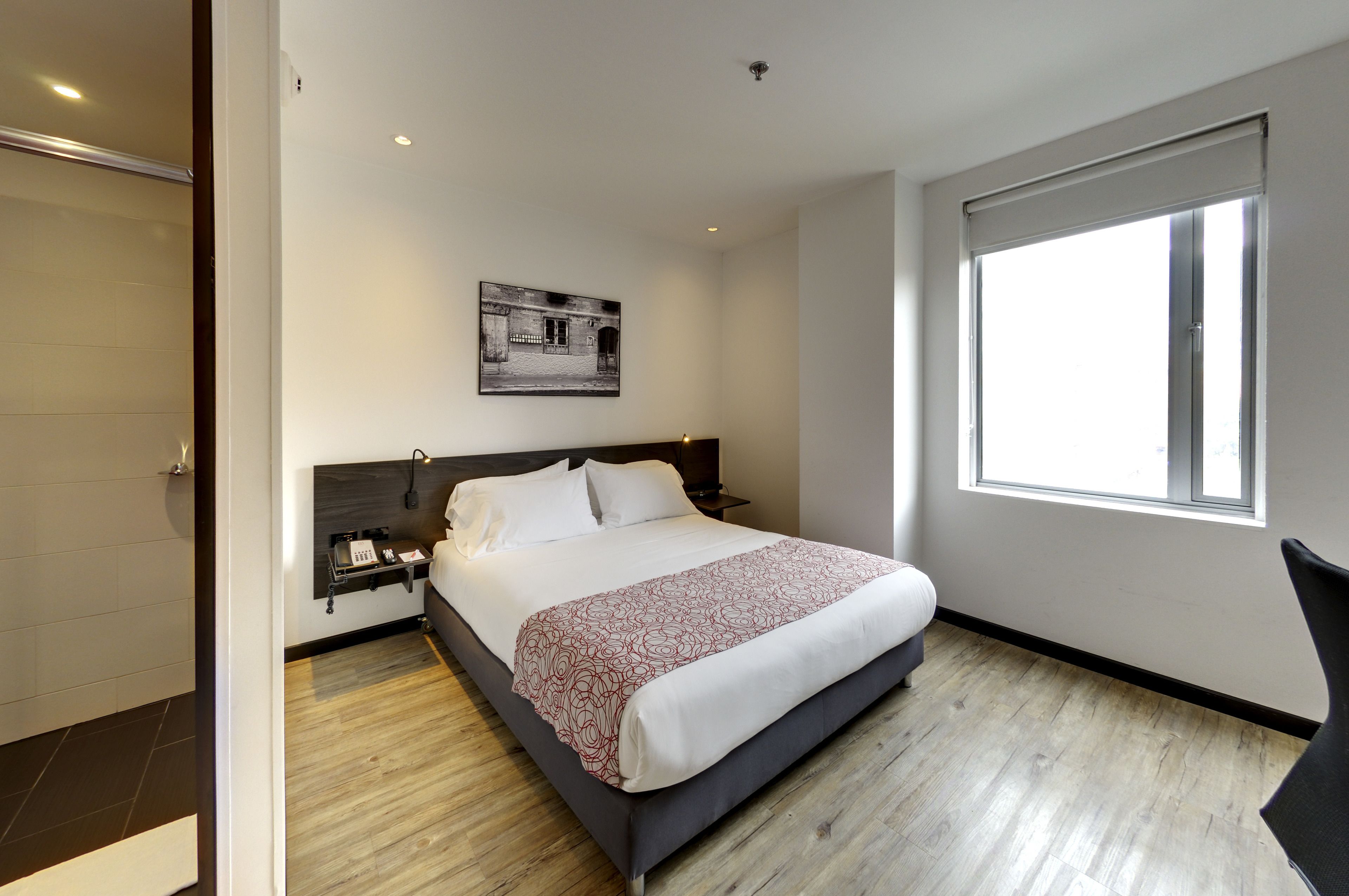 A modern hotel room with a large bed, a bedside table, a wall-mounted phone, and a window with a view.