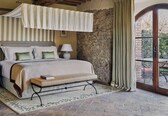 A bedroom with a canopy bed, two bedside tables with lamps, a bench, a rug, and a stone wall.