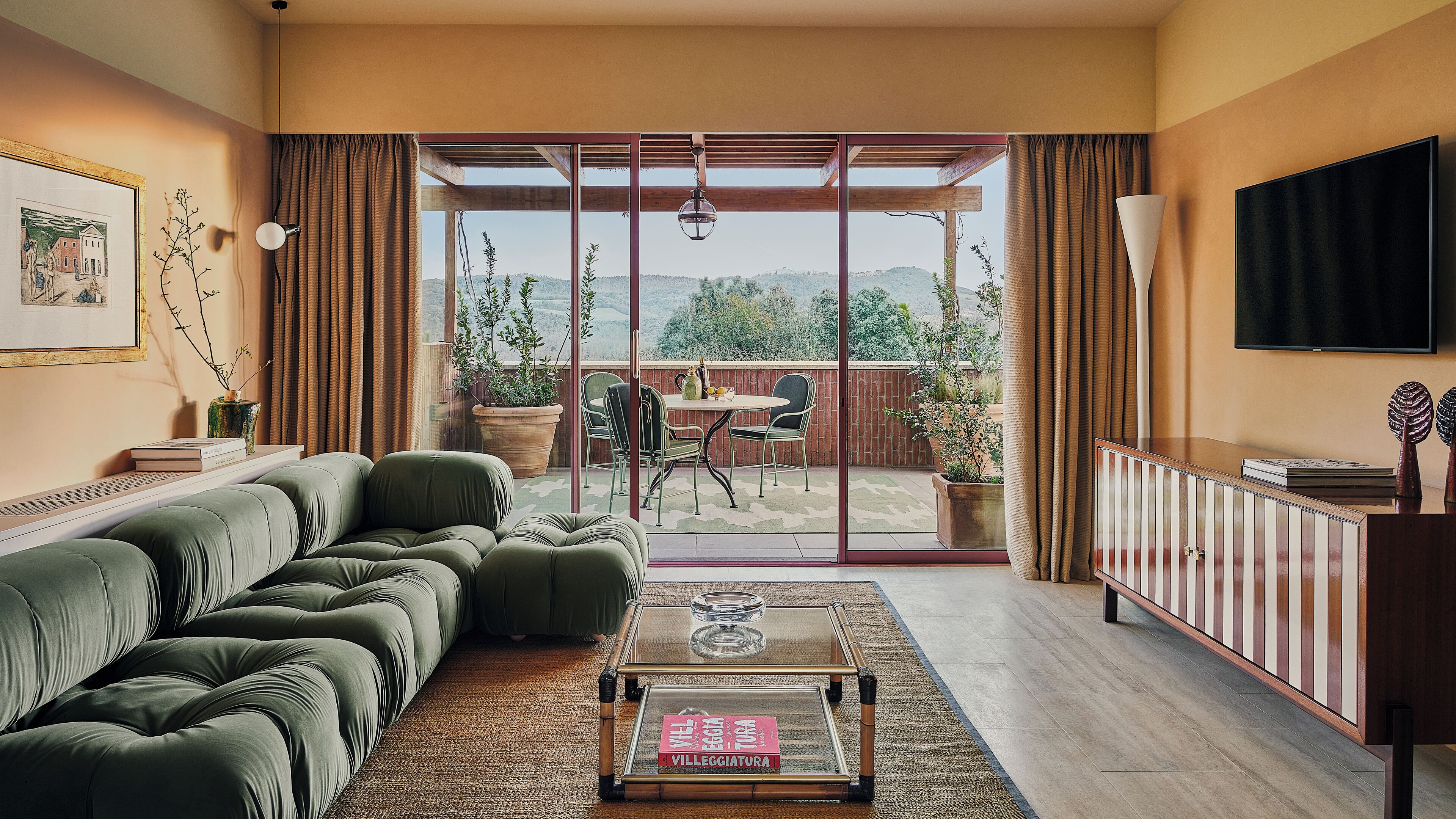 Un salon avec un canapé, une table basse sur laquelle repose un livre, un téléviseur et une vue sur l’extérieur grâce à de grandes portes vitrées.