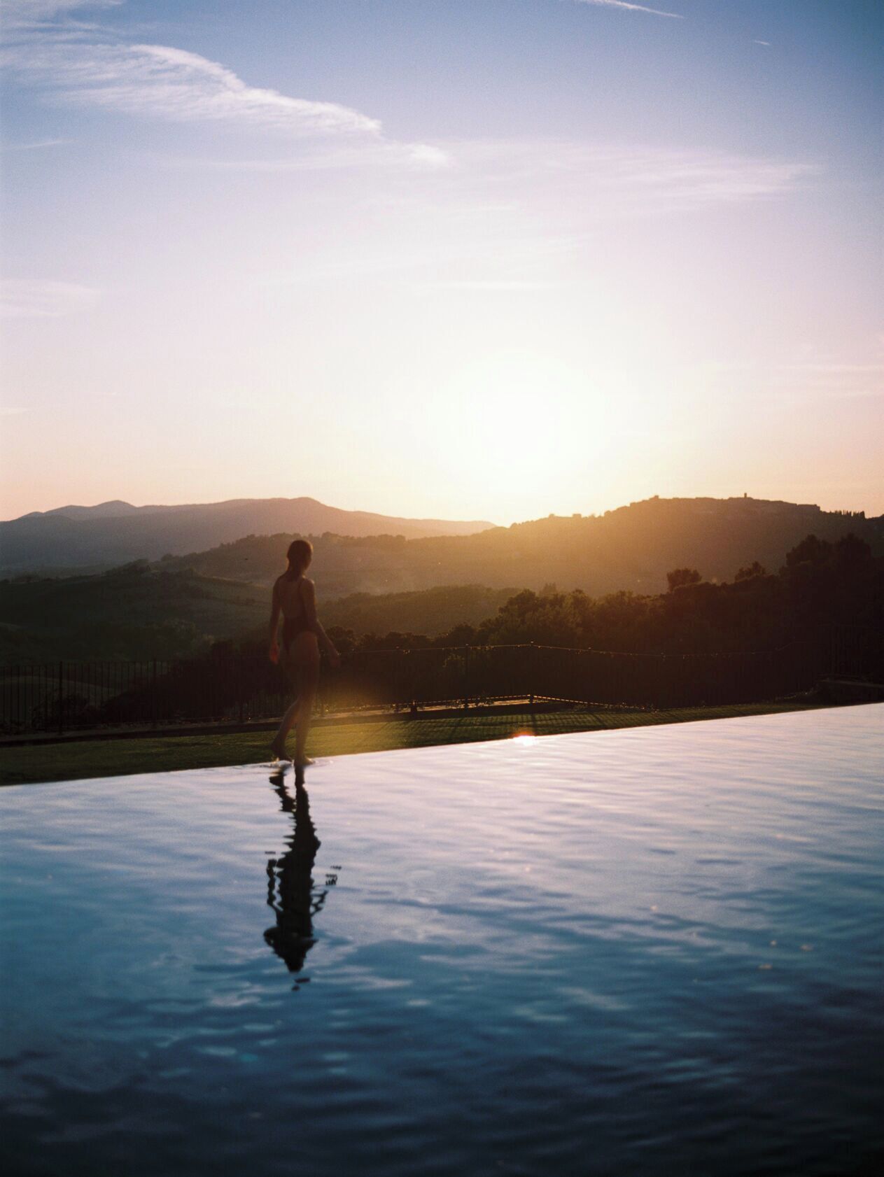 Une personne se tient au bord d’un bassin dont la surface est réfléchissante, avec une vue sur des collines lointaines et un coucher de soleil.