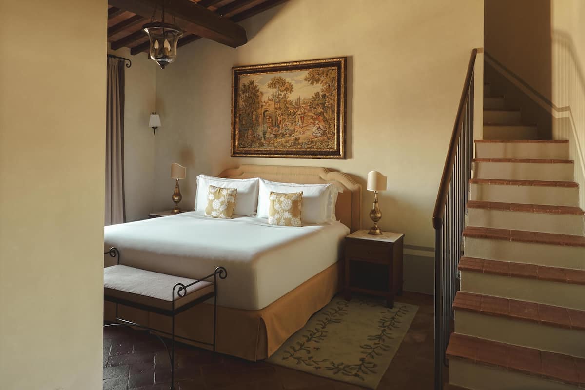 A bedroom with a bed, bedside tables, a painting, a staircase, and a bench.