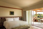 A hotel room with a large bed, a bedside table with a lamp, a small table with chairs, and a sliding glass door to an outdoor area with plants.