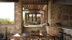 Patio esterno con parete in pietra, tavolo e sedie di legno, piante in vaso e camino in pietra.