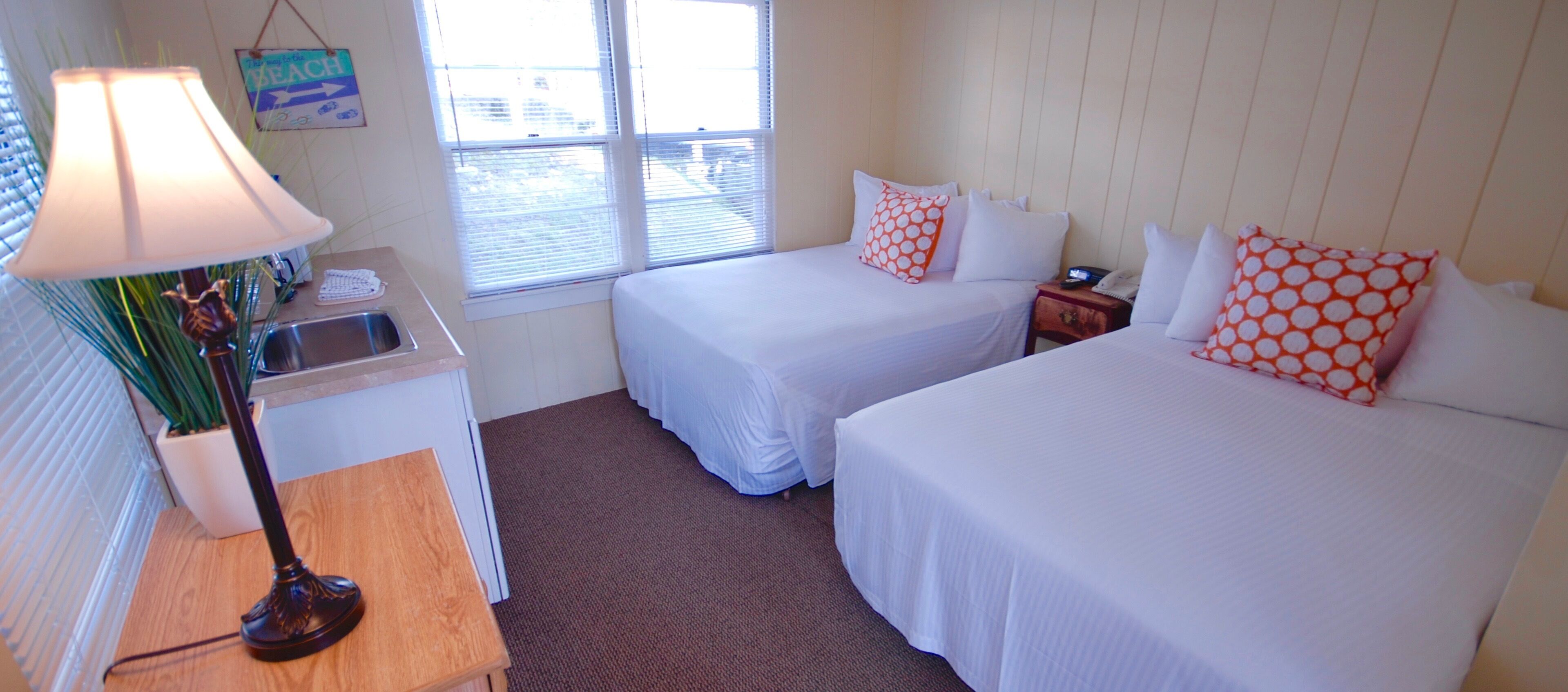 Habitación de hotel con dos camas, un escritorio con lámpara, un lavamanos pequeño y una ventana con persianas.